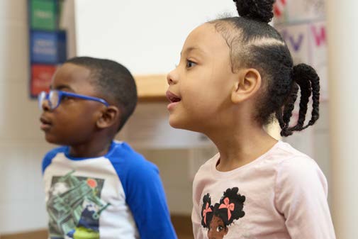 Two young children in a classroom, attentively looking to the left.